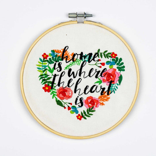Embroidery on a hoop with colorful floral design and text 'home is where the heart is' on a white background